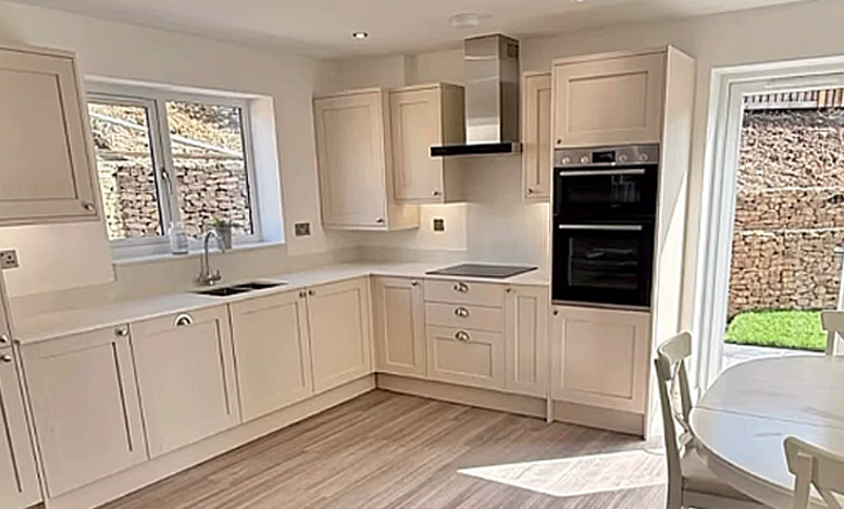 Modern cream-coloured fitted kitchen with an L-shaped layout, integrated double oven, electric hob, extractor hood and sink beneath a window.