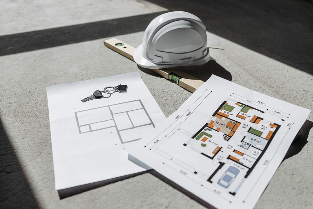 Architectural floor plans and building drawings laid out on a table beside a white hard hat, spirit level and set of keys.